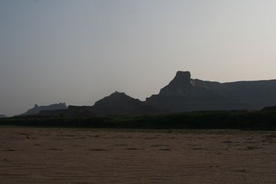 A stepped butte and long mesa rim stand in near-silhouette against a hazy dusk sky, with dark riverside brush and a wide rippled sand flat in the foreground.