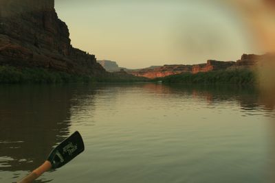 View forward from a boat down a glassy river reflecting a pale gold sky, an oar blade dipping in at lower left and low sunlit red cliffs closing the canyon in the hazy distance.
