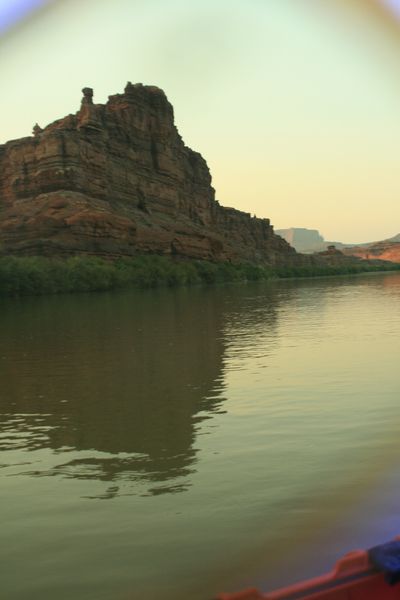 Vertical frame from a boat looking at a layered red cliff capped by a small knob of rock, mirrored in glassy green-gold water, with the orange edge of an inflatable visible at the lower right corner.