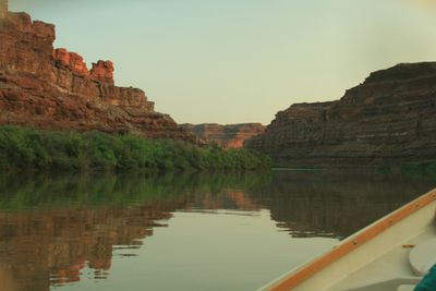 View past the varnished wooden gunwale of a dory down a glassy river, sunlit red spires standing above a dark layered wall on the left and the canyon bending away between banks of green brush.