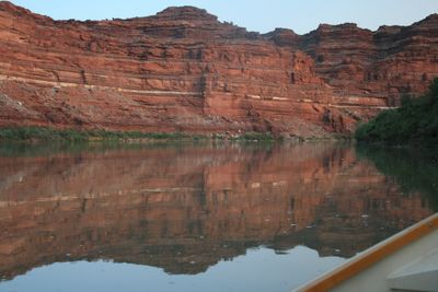 A red canyon wall banded with a bright white layer and skirted by rubble slopes stands mirrored in perfectly still water, with the wooden gunwale of a dory across the lower right corner.