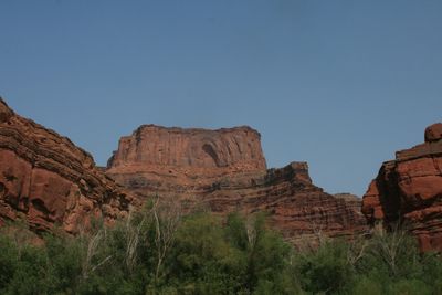 A broad red sandstone butte with a dark arched recess in its face rises above a slope of layered ledges and a screen of green riverside trees, under a deep blue midday sky.