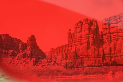 Castellated sandstone cliffs and detached pinnacles above a brushy riverbank, the entire frame washed out by a heavy red light leak that obliterates the sky and flattens all colour.