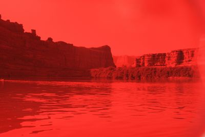 A wide flatwater bend below layered cliffs and a thick riverside brush line, with a small boat just visible at the far left edge of the water — the whole frame saturated red by a light leak.