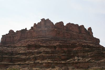 Looking up at a large stepped butte of red and gray rock layers topped by rounded knobs, its lower slopes covered in rubble, under a pale hazy sky.
