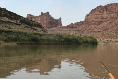 From a boat on a wide olive-green river, a grassy bench and brush line lead back to layered red cliffs with a castle-like formation on the skyline, an oar blade entering the water at lower right.