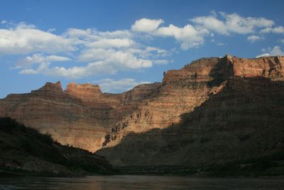 Sunlit layered cliffs and rock towers rise beyond a dark shadowed foreground ridge, with a strip of river at the bottom of the frame and cumulus clouds across a deep blue sky.