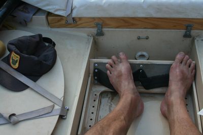 Looking straight down into the footwell of a dory at a pair of bare feet braced against a strapped foot bar, with a gray ball cap, cam straps and a hatch lid on the deck alongside.