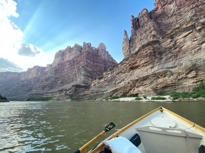 Wide phone-camera view from the bow of a wooden dory: sunlit layered walls and clustered rock towers rise on both sides above a green brush line and pale sand bank, with an oar shipped across the foreground and sunbeams fanning across a blue sky.