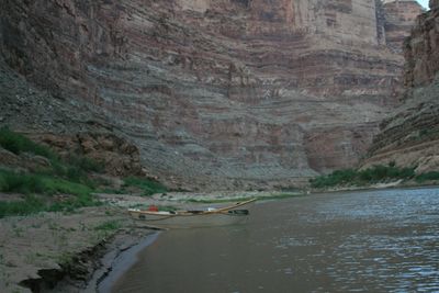 A white wooden dory lies beached on a sand bar at the water's edge below an enormous wall of gray and tan rock layers, in flat blue dusk light with green brush along the shoreline.