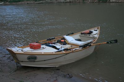A white wooden dory rigged with wooden oars, an orange dry box and an orange life jacket sits beached on wet mud and sand at the river's edge in flat dusk light.