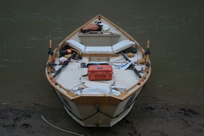 Bow-on overhead view into a beached white wooden dory showing shipped oars, padded seats, an orange hard case on the deck, a folded map, a metal cup and a yellow bow line running to shore.