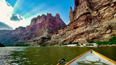 From the bow of a white dory on flat water in lower Cataract Canyon, red sandstone spires and layered cliffs rise above a white sand beach fringed with tamarisk, sun rays breaking through cloud.