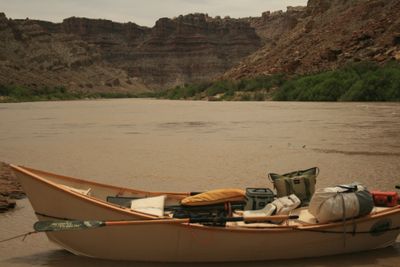 A loaded wooden dory pulled up on a muddy bank of the silt-brown Colorado River, rigged with dry bags, coolers, an orange hard case and oars, with layered canyon walls and green riverbank brush across the water under a hazy sky.