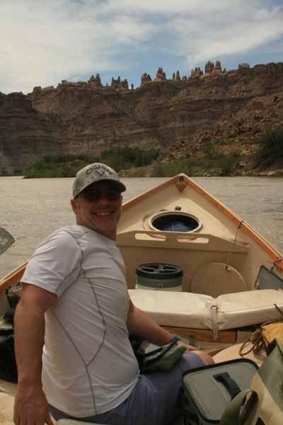 A smiling crew member in a white long-sleeve sun shirt, ball cap and sunglasses sits in the wooden dory on the silt-brown river, the boat's hatch, bucket and dry bags around them and sandstone spires on the horizon.