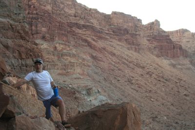A crew member in a white t-shirt and ball cap stands among boulders on a steep talus slope holding a blue insulated cup, mouth open mid-word, below tall banded cliffs under a flat white sky.