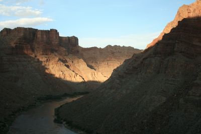 Late sun lights a wedge of banded cliff and slope above the shadowed inner canyon while the silt-brown river runs through deep shade at the bottom of Cataract Canyon.