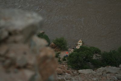Looking down from high on the canyon wall past a blurred foreground rock at the wooden dory tied to a small sand beach, camp gear spread among green riverside brush, with the silt-brown river filling the rest of the frame.