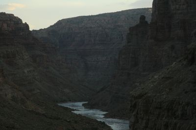 The silt-brown river threading the bottom of a deep, shadowed canyon at dusk, layered walls stepping back to a distant mesa rim under a pale sky.