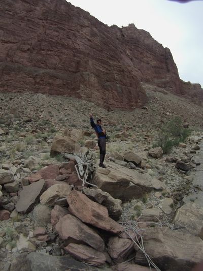 A crew member in a blue drytop and black pants stands with arms spread on a boulder-strewn bench below a tall banded canyon wall, bleached driftwood and brush scattered around them.