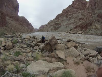 A crew member in a blue drytop stands among huge fractured sandstone blocks on the bank while the silt-brown Colorado runs fast past the debris field, layered canyon walls rising on both sides under an overcast sky.