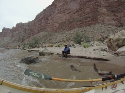 Seen over the wooden dory's gunwale and shipped oar, a crew member in a blue drytop sits on a sandstone ledge at the edge of the silt-brown river on a sandy bench, driftwood and brush behind them below a tall red canyon wall.