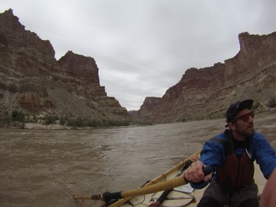 The boatman, in a blue drytop and red life vest, works the oars of the wooden dory on the silt-brown Colorado with a river map open on the deck, layered canyon walls rising on both sides under an overcast sky.