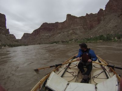 From the stern of the wooden dory, the boatman rows down the silt-brown Colorado past layered canyon walls and a distant mesa, the boat's hatch lids, cam straps and river map spread across the deck.