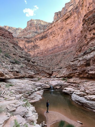 A hiker stands at the edge of a clear pool in canyon bedrock with a small cascade feeding it, beneath a tall varnished red sandstone wall.