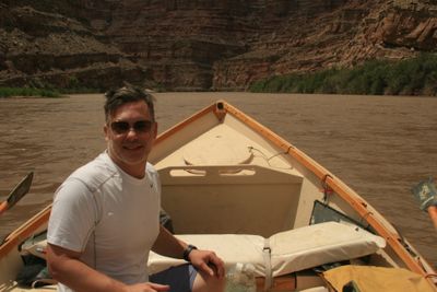 A crew member in a white shirt and sunglasses sits in the bow of a wooden dory on muddy brown water, with layered red canyon walls behind.