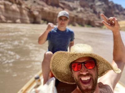 Two crew members mug for a wide-angle selfie on the boat, one grinning in a straw hat and red mirrored sunglasses, muddy river and red canyon wall behind.