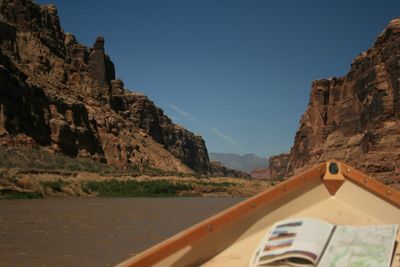 Looking downstream over the bow of a wooden dory with an open river guide map on the deck, canyon walls opening toward distant mountains on the horizon.
