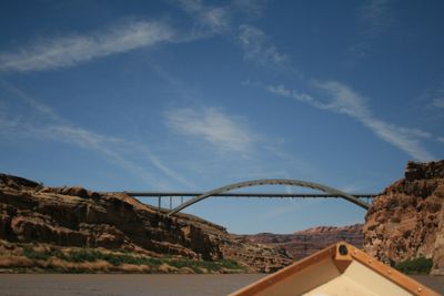 A steel arch highway bridge spans the canyon ahead, seen over the bow of a wooden dory under a cirrus-streaked blue sky.