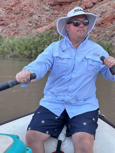 A crew member in a wide-brimmed sun hat and light blue fishing shirt pulls on the oars of a river boat, with muddy brown water and a red talus slope behind.