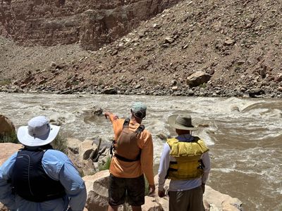 Three crew members in life jackets and sun hats stand on a rocky bank scouting a large brown rapid, one pointing at the current line.