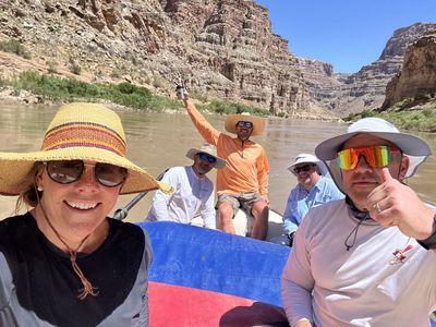 Five crew members in sun hats and sunglasses pose for a selfie on a rigged raft on flat brown water, with layered canyon walls opening downstream behind them.
