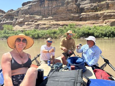 Four crew members relax on a rigged raft holding drinks, with oars, dry bags and a blue bucket aboard and a tan cliff band above green riverside brush.