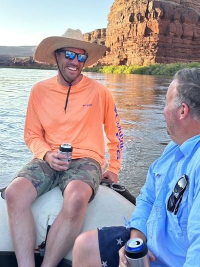 A crew member in an orange sun shirt and straw hat laughs while holding a drink on the raft tube, with warm low light on a red sandstone wall across glassy water.