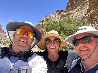 Three crew members in life jackets crowd into a close selfie, one with an action camera mounted on his shoulder strap, with a rocky slope and a green tree behind.