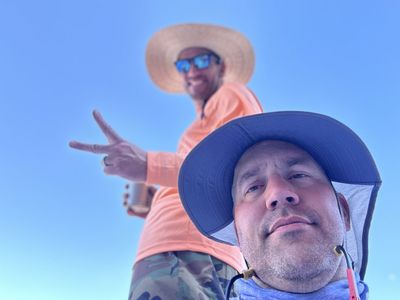 A low-angle selfie against a cloudless blue sky: one crew member in a navy sun hat in the foreground, another behind in an orange shirt flashing a peace sign and holding a can.