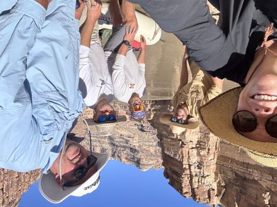 Five crew members gather for a selfie on a rigged raft against tall pale sandstone walls and buttes, with flat silt-brown water behind them.