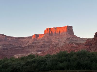 Warm low-angle sunlight strikes only the top of a red sandstone butte above shadowed, stepped canyon benches, with a dark band of riverside trees across the foreground.
