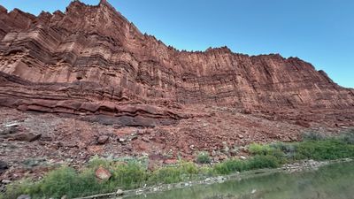 A tall banded red sandstone cliff rises above a rubble slope and a green scrub-lined riverbank, photographed from water level with slight motion blur under a clear blue sky.