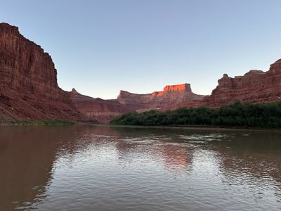 Warm low sunlight lights a distant butte and rim above a bend of the Colorado River while the near walls stand in shadow; the brown water carries a rippled reflection of the lit rock.