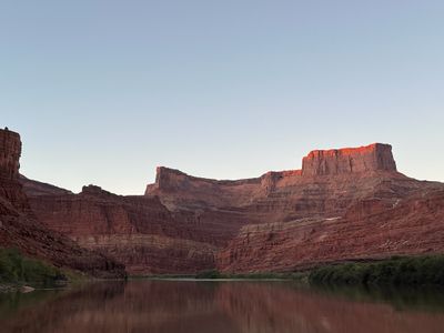 A thin band of warm sunlight burns along the highest cliffs and a butte cap above stepped red benches, with the still brown river reflecting the wall in the foreground.