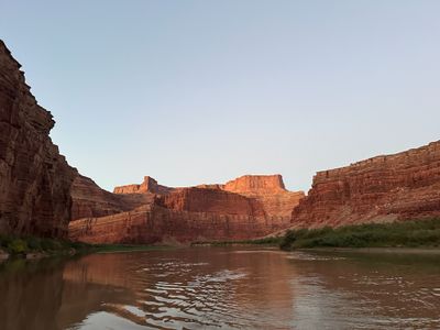 Looking downstream on the Colorado between shadowed sandstone walls toward a warm-lit butte and rim, with rippled brown water filling the lower half of the frame.