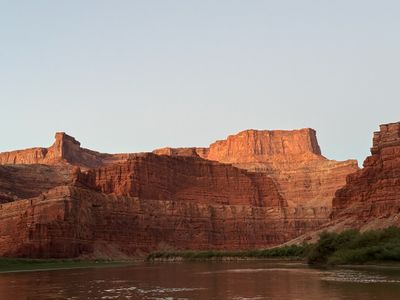 A broad warm-lit sandstone wall and butte rise in layered ledges above a narrow green strip of riverside brush and the brown Colorado River.