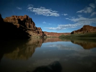 A moonlit night on the river: a deep blue sky with lit clouds and a few stars over dark canyon walls, mirrored almost perfectly in glassy water.
