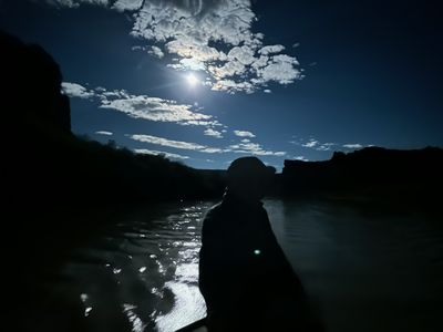 A crew member sits silhouetted at the oars against a moonlit river; the moon burns through broken clouds overhead and lays a bright glitter path on the water, and a small green glow-stick shows on the rower's chest.