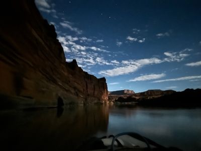 From the boat at night: a moonlit canyon wall glows warm on river left under a starry deep blue sky with streaked clouds, and the still water reflects the lit rock; a pale raft frame edges into the bottom of the frame.